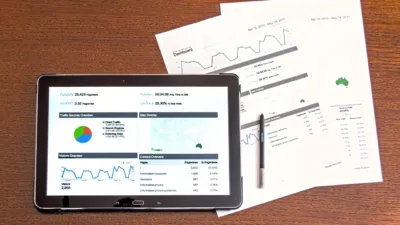 Tablet showing an analytics dashboard beside printed business reports, illustrating how NetSuite SuiteAgents monitor finance and operations workflows