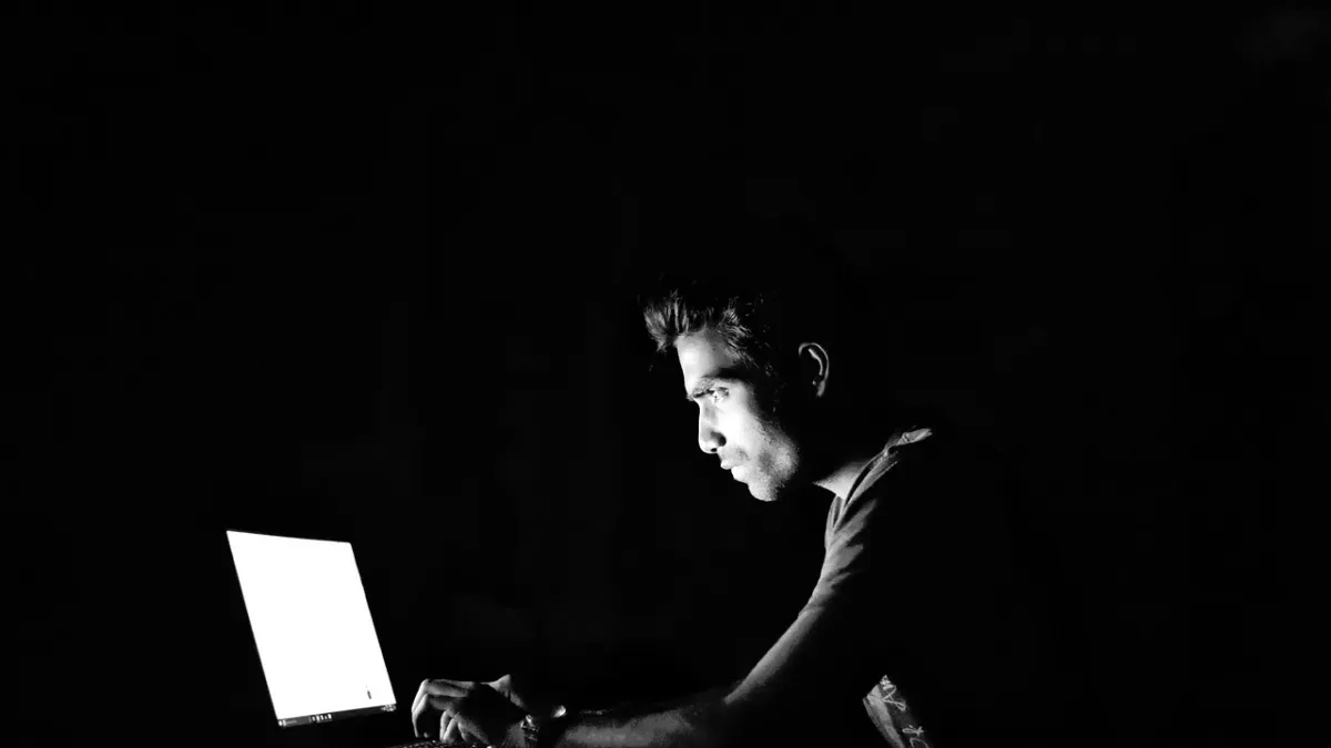 Hooded hacker typing on a laptop in a dark room, illustrating the AI agent cyberattack that breached 100+ company websites