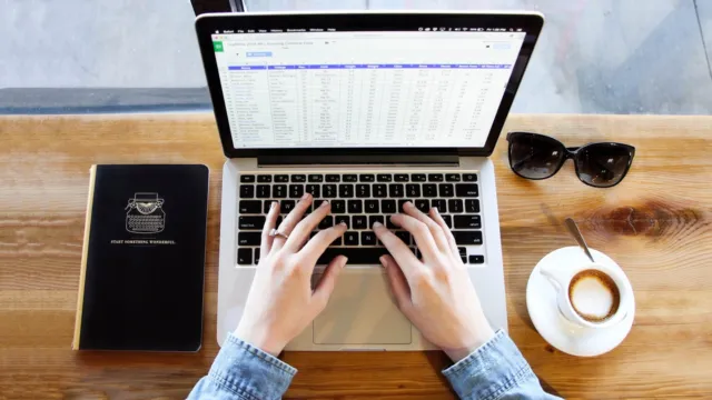 Top view of hands typing on a laptop with coffee and a spreadsheet on a wooden desk, illustrating AI formulas in Excel and Google Sheets